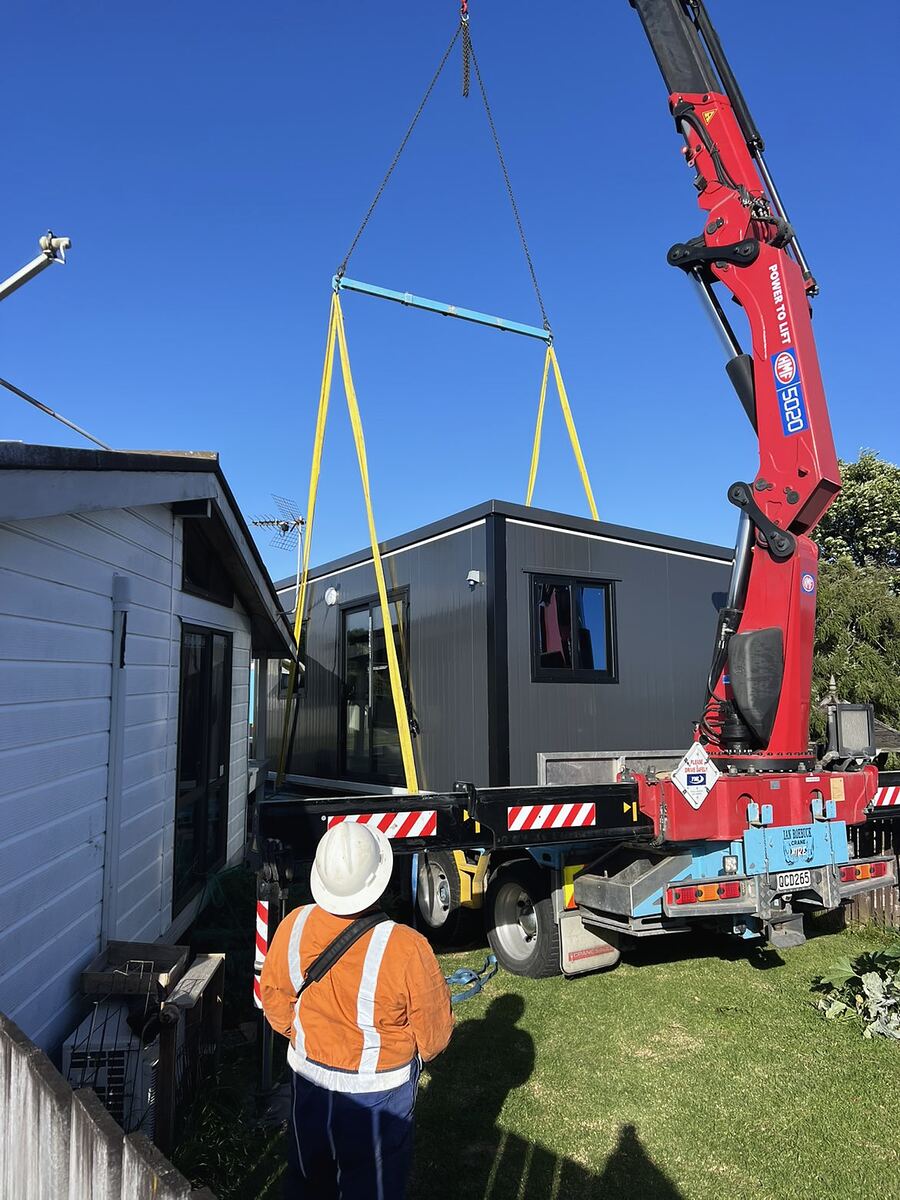 A hiab crane lifting a cabin into a backyard beside a house